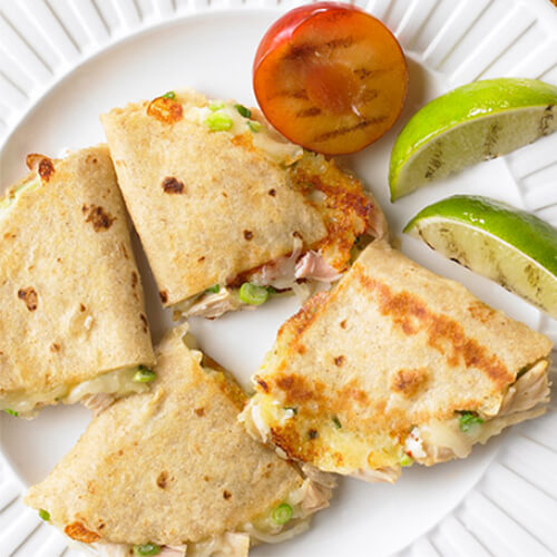 Quesadillas filled with spicy Monterey jack cheese, roasted turkey, and fresh green onions, served with a side of grilled tomatoes and lime wedges, on a white plate with an orange tablecloth.