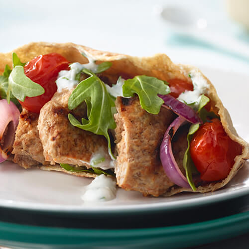 Spiced turkey meatballs covered with in vegetables, an herbed yogurt, all stuffed in a pita, on a white plate on a teal table.