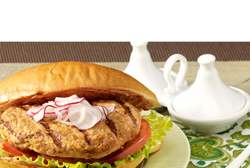 A turkey burger spiced up with harissa, and served with lettuce, tomato, and onion, topped with a sweet radish mayo, on a mint green plate atop a green floral napkin.