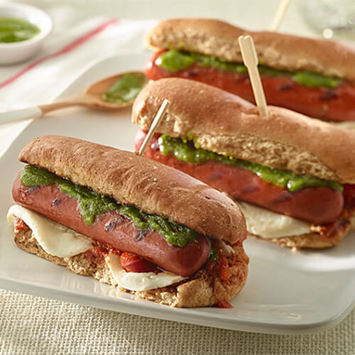 Franks, melted mozzarella cheese, roasted bell peppers, pesto and marinara, all served in a hot dog bun on a white platter.