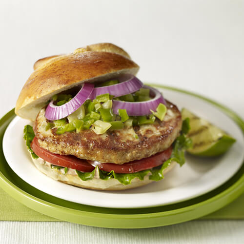 A juicy turkey burger made with pepper jack,and topped with onions, lettuce, tomato, and chili mayo, served on a green painted plate garnished with grilled lime.