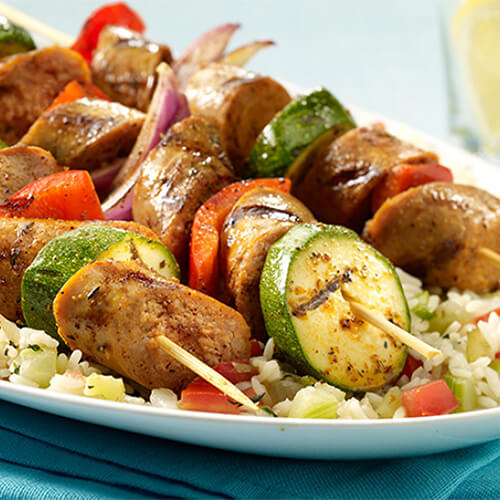 A New Orleans inspired kabob made with juicy hot Italian sausage and grilled veggies with Cajun seasoning, served with veggie rice in a white plate on a cyan cloth.