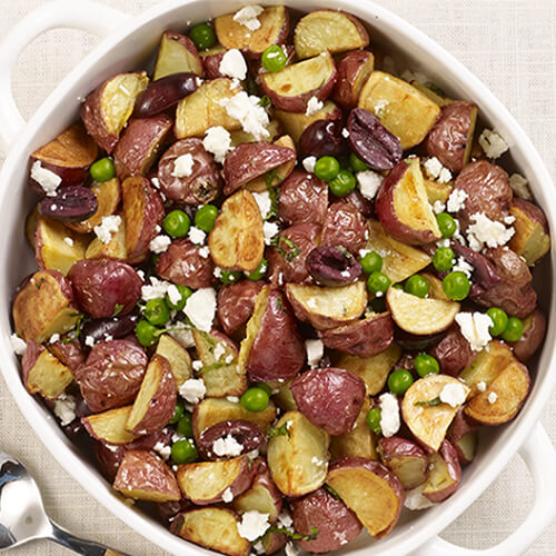 A bowl of potatoes featuring tossed feta cheese, Kalamata olives and mint, served on a white plate atop a linen tablecloth.