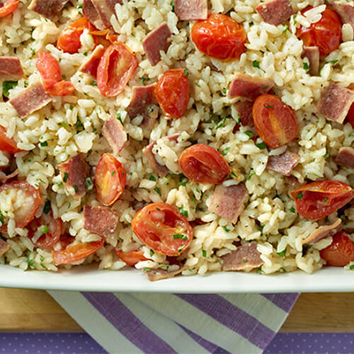 A hearty portion of grape tomatoes, bacon, and risotto rice in a green tray on a wooden board.