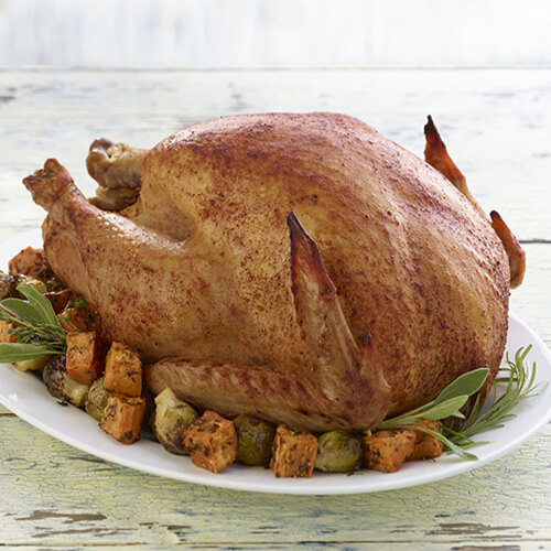 A gluten-free turkey on top of vegetables, sweet potatoes and herbs on a white plate with a wooden background.