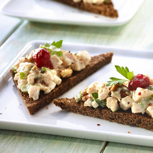 Slices of pumpernickel bread, topped with a mixture of turkey, mayonaise, spices, and pecans, topped with cranberry sauce, served on a white plate with a side of cranberry sauce.