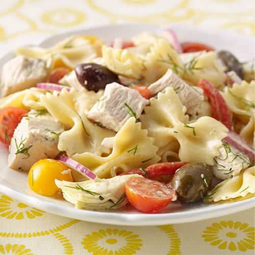 Bow tie pasta tossed with artichokes, cherry tomatoes, lean turkey breast and fresh dill, served in a white plate on a yellow patterned tablecloth.