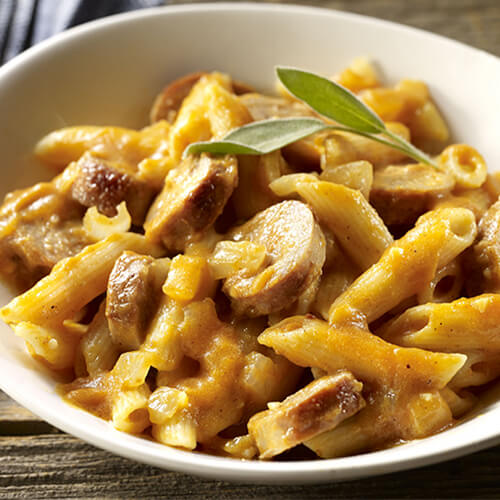 A bowl of penne pasta made with sweet Italian sausage, and a sauce made from pumpkin, white wine and, chicken broth with a wooden background with a blue napkin and a fork.