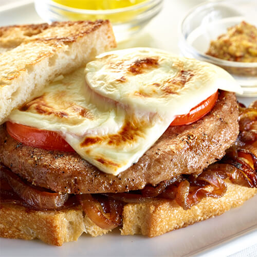 Toasty bread topped with caramelized onions, a juicy turkey burger patty tomato, and Swiss cheese, served with a side of grain mustard, on a white plate and tablecloth.