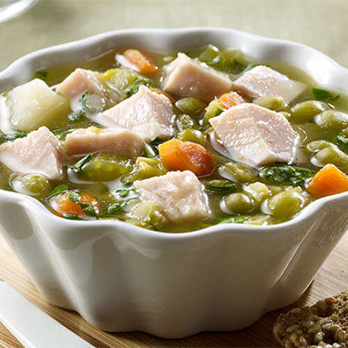 A hearty portion of split peas, potato, celery carrot and herbs, filled with smoked turkey breast, served in a white bowl atop a wooden board.