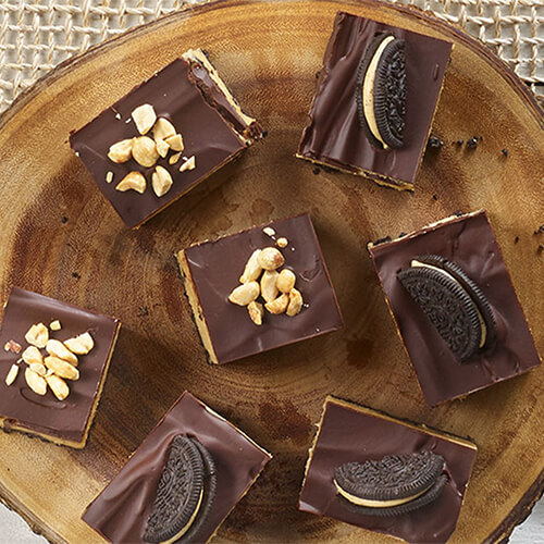 Peanut butter and chocolate cheesecake bars garnished with nuts on a wood plate.