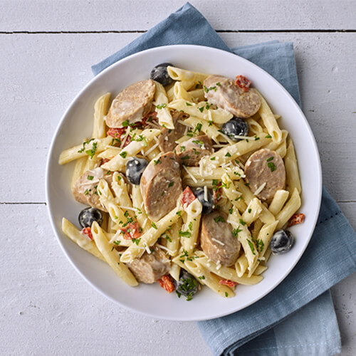 A heaping portion of penne pasta, topped with creamy Alfredo sauce, ripe olives and tomatoes, spicy Italian turkey sausage, and parmesan cheese, on a blue napkin and a white wooden table.