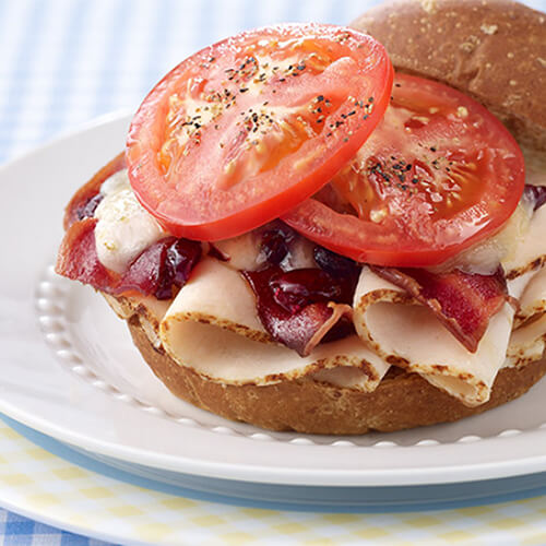Crispy bacon, fresh-sliced deli turkey, cranberries, melted pepper jack and fresh tomato on a whole wheat bun, in a patterned plate on a blue checkered tablecloth.