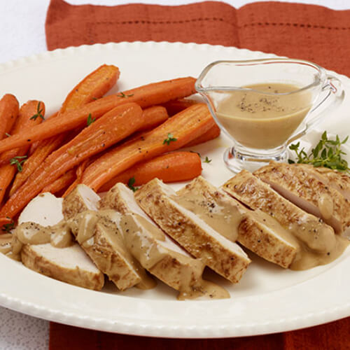 Glazed turkey tenderloin drizzled with peppercorn sauce, with a side of more peppercorn sauce and buttery baked carrots, on a white plate, an orange napkin, and on a white linen tablecloth.