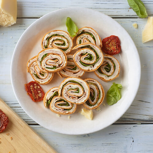 A white plate of pesto turkey pinwheels made of JENNIE-O® Deli-Sliced Turkey, lettuce, and pesto. Next to sun dried tomatoes and fresh picked basil leaves.