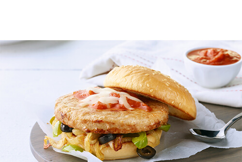 A turkey burger topped with peppers, olives, pizza sauce, and cheese, served on parchment paper atop a wooden plate.