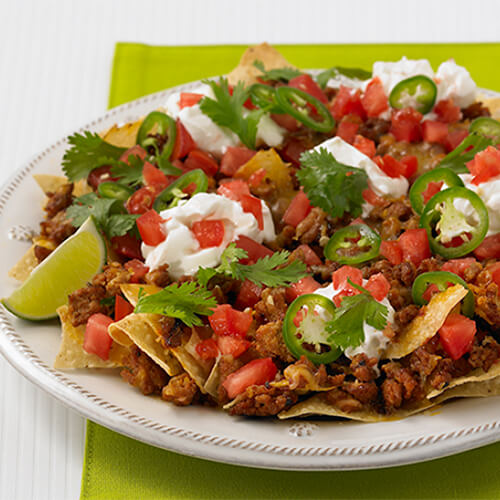 A hearty portion of tortilla chips and ground turkey, topped with jalapeno, cilantro, tomato, cheese, and sour cream, garnished with a lime wedge, served with a side of salsa on a white plate and table.