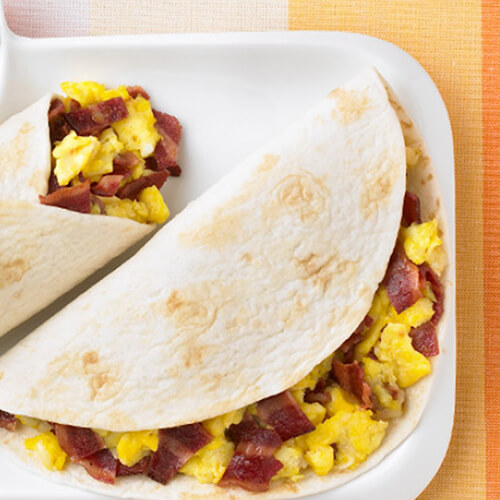 2 hearty breakfast burritos, stuffed with scrambled eggs, turkey bacon and colby cheese in a warm flour tortilla, garnished with orange slices and a sprig of cilantro, and served with salsa, in a white tray, on a vibrant tablecloth.