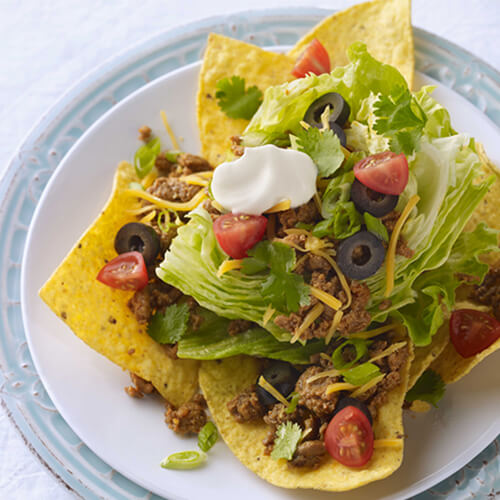 A great amount of ground turkey, vegetables, cilantro, cheese, and sour cream served on chips, on a painted plate.