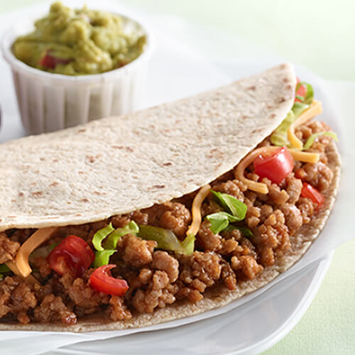 A hearty portion of ground turkey, lettuce, tomato, and avocado stuffed inside a warm tortilla on a white plate, served with a side of grapes and guacamole.