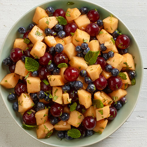 Cantaloupe, grapes and blueberries mixed into a bowl, and served with a dressing made from sugar, ginger, and honey, served in a green bowl atop a wooden table.