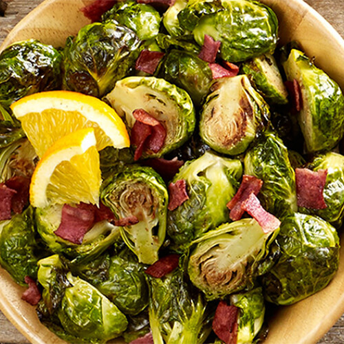 Roasted brussels sprouts tossed with bacon, mustard, and honey, garnished with a lemon slave in a wooden bowl atop a table.