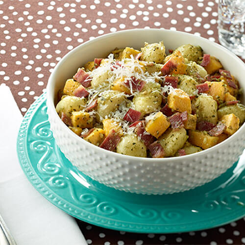Pillowy gnocchi and crispy turkey bacon with roasted butternut squash, garnished with parmesan cheese in a white studded bowl atop a blue patterned plate.