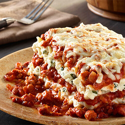 A hearty classic lasagna filled with ground turkey, basil, pasta sauce, ricotta, asiago, and mozzarella cheeses, on a wooden plate and table.