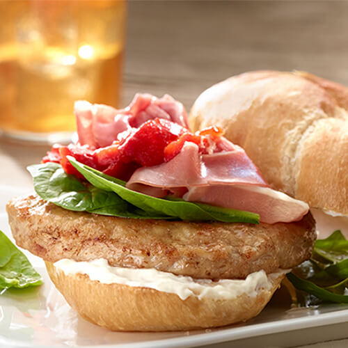 A turkey burger topped with red pepper, arugula, prosciutto and mayonaise, served on a white plate.