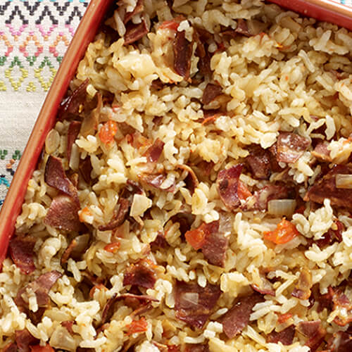 A hearty risotto full of turkey bacon served in an orange baking dish, and served with a homemade tomato granita in a blue bowl, both atop a painted tablecloth.