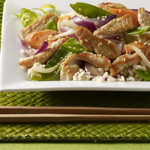 A hearty stir-fry featuring turkey tenderloins simmered with carrots, bell peppers, onions, fresh ginger and pea pods, served on rice with a side of soy sauce, on a green place mat with chopsticks.