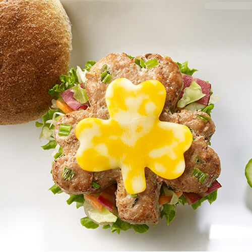 A shamrock-shaped, bacon-filled, cabbage kraut topped with coleslaw and cutout shamrock cheese, served on a whole wheat bun on a white tray with a side of pickles.