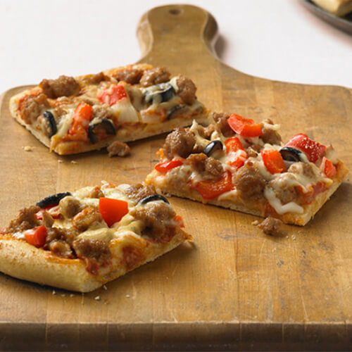 3 squares of Sicilian pizza topped with turkey, red bell pepper and olives on a wooden pizza board, with a pink background.