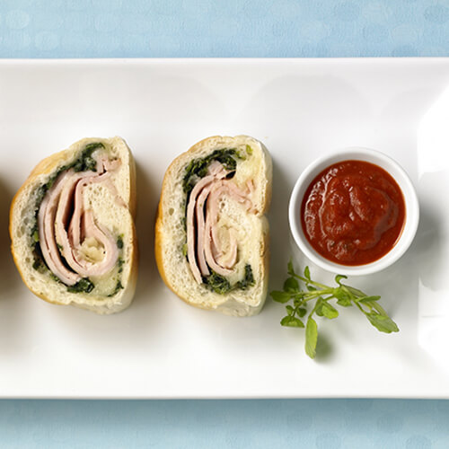 3 Strombolis baked into a flaky crust filled with spinach and provolone with a side of marinara to dip, on a white platter in a blue tablecloth.