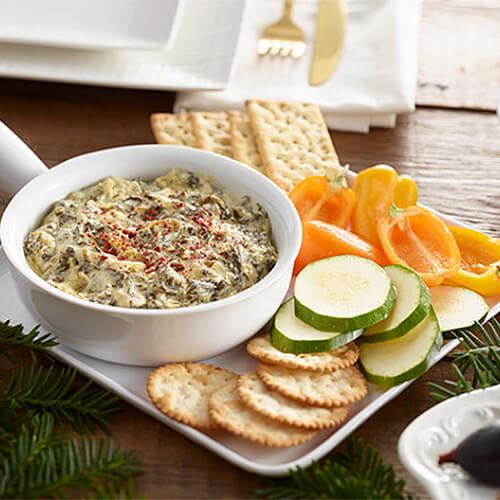 Spinach dip in a white bowl, on a plate with crackers and vegetables.