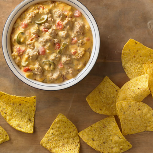 A blue lined bowl filled with jalapeno peppers, JENNIE-O® ground turkey, turkey sausage, vegetables, and a variety of cheeses. with a side of corn chips placed on parchment paper.