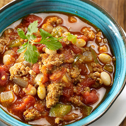 Spiced turkey chili made with tomatoes, beans, salsa and quinoa, served in a blue bowl atop a white plate on a wooden table.