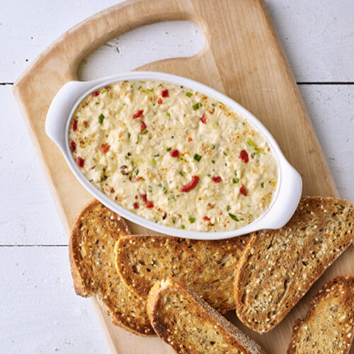 A white serving bowl of melted smoked gouda cheese and turkey dip with crunchy bread on a wooden cutting board.