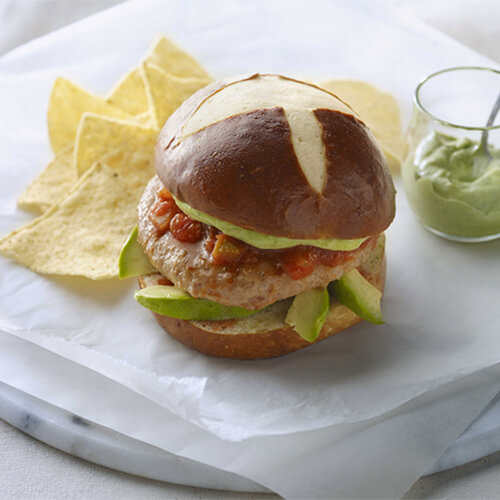 JENNIE-O® turkey burger patty, pepper jack cheese, cilantro pesto, avocado slices, and CHI-CHI'S® salsa on a pretzel bun, served on a marble plate with tortilla chips and more cilantro pesto,