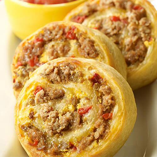 3 pinwheels made from cheese, egg, and lean turkey sausage encased in a flaky pizza crust, served on a white plate with a side of marinara sauce on a colorful tablecloth.