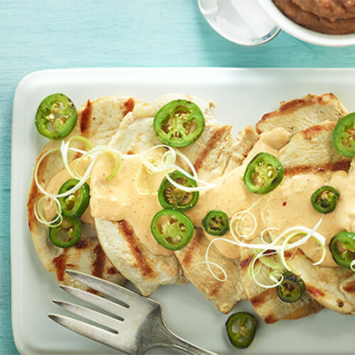 Tender grilled turkey breast topped with a portion of jalapenos and a large portion of spicy cheese sauce, served on a white plate with a side of beans, on a blue tablecloth.