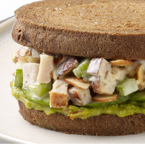 A hearty turkey salad sandwich topped with spicy guacamole, served on rye bread on a white plate.