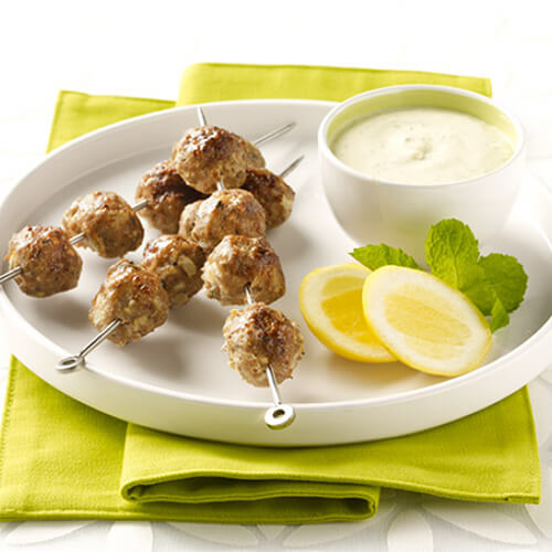 3 mouth-watering turkey kabobs are baked with garlic, mint and coriander served with a side of tangy yogurt sauce, garnished with mint and lemon slices, served on a white plate on a green napkin.