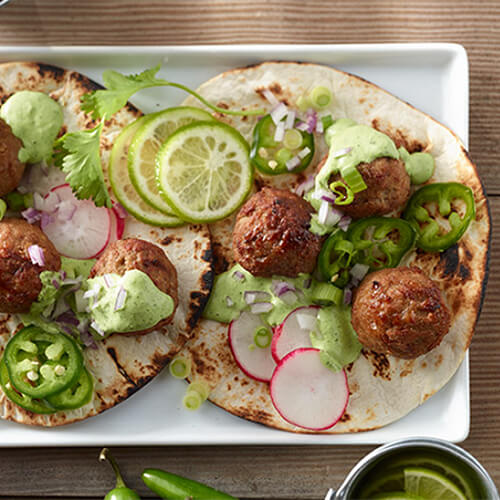 Charred soft tortillas, topped with crunchy radishes, savory turkey, limes, and a spicy cilantro dressing, served on a white platter atop a wooden table.