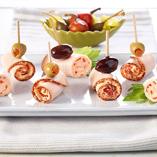 Spicy roll-ups filled with fresh jalapenos blend with cool cream cheese, sharp cheddar and lean turkey breasts, topped with olives, served on a white tray.