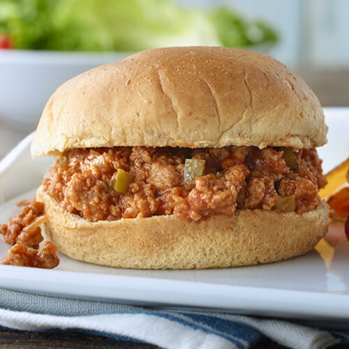 Turkey sloppy joe meat made with yellow bell pepper in a thick hamburger bun next to side of chips on a white plate.