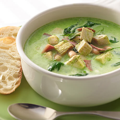 A creamy spinach soup filled with beans, bacon, and turkey, served in a white soup bowl, with a side of bread on a green plate.