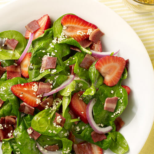 A fruity, filling, salad filled with Fresh strawberries, turkey bacon, and baby spinach, served with a vinaigrette, in a white plate on a yellow lined tablecloth.