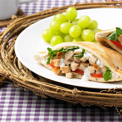 A pita pocket with turkey, tarragon leaves and tomato, drizzled with ranch dressing with a side of grapes, served in a white plate and a woven bowl, on a purple checkered tablecloth.