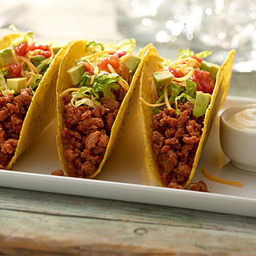 Simple tacos made with round turkey, shredded cheese and veggies, with a side of sour cream and salsa, on a white platter.